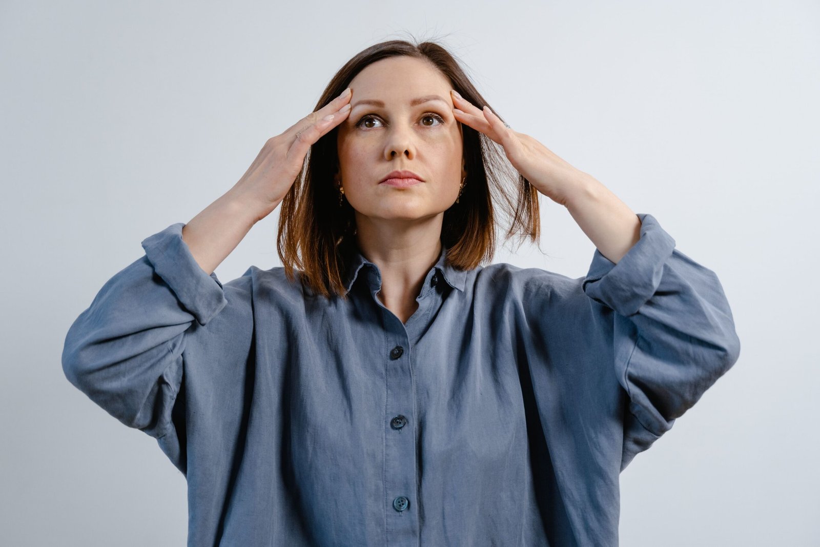 Woman in gray shirt with hands on head, looking thoughtfully upward indoors.